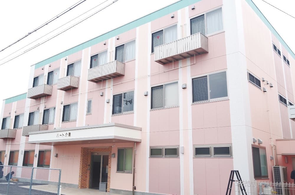 ピンク色の3階建て介護施設の建物外観。複数の窓とバルコニーが見える。
